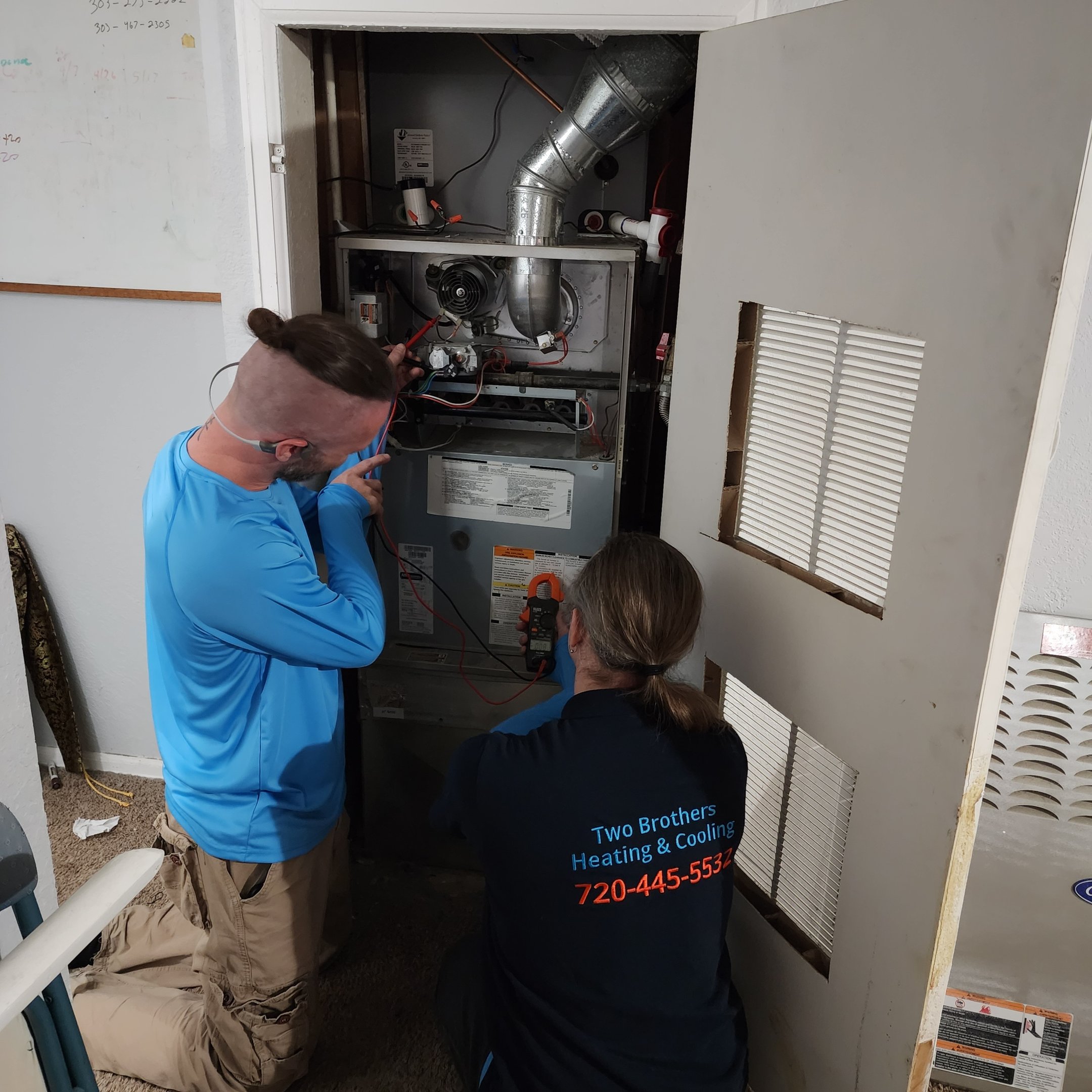 Two Brothers technicians at an open furnace closet, one holding meter probes on the gas-valve wiring while the other reads a clamp meter