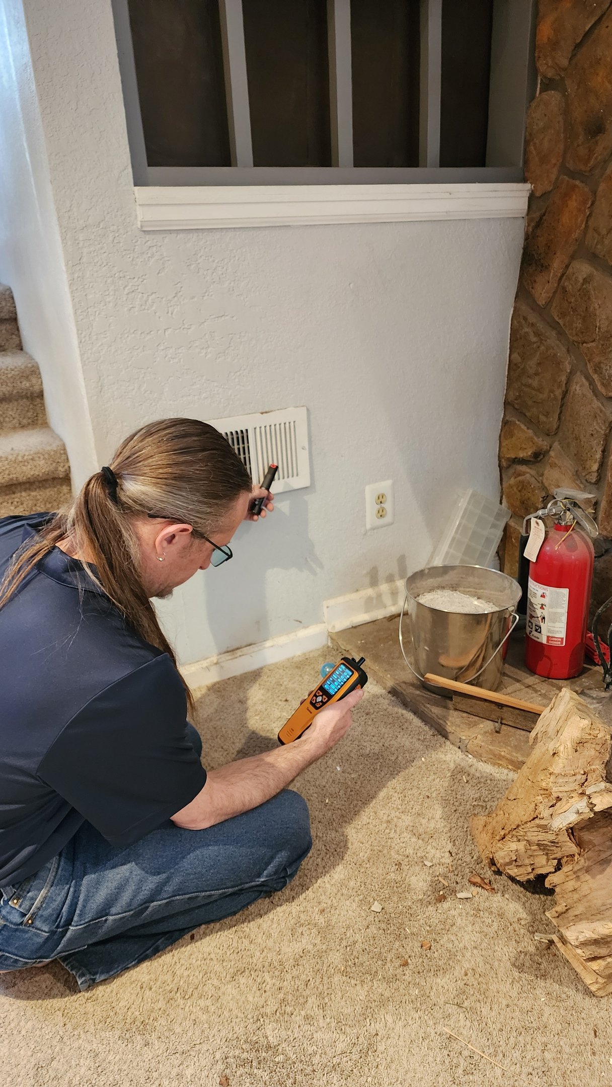 Two Brothers technician kneeling at a floor-level return vent with a handheld air quality meter